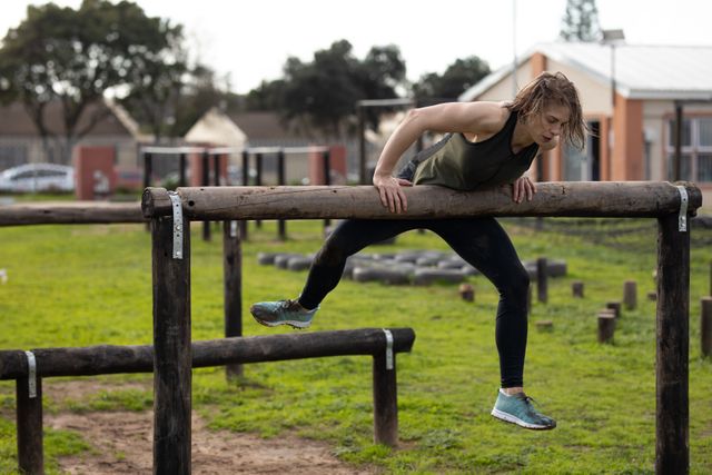 Front view of a Caucasian woman wearing sports clothes climbing over a tall wooden hurdle at an outdoor gym during a bootcamp training session, with other outdoor gym equipment in the background