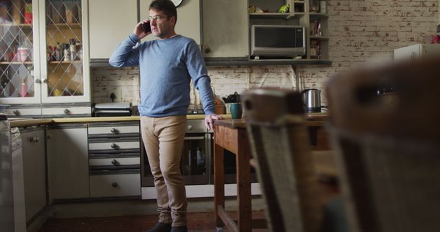 A man wearing a blue sweater and light brown pants is talking on the phone while standing in a cozy kitchen with rustic decor. The kitchen features brick walls, wooden furniture, and traditional cabinetry. Useful for advertisements depicting home life, telecommunication services, or casual daily routines.