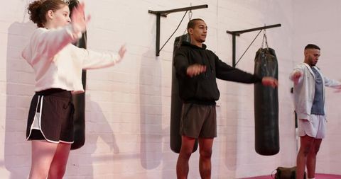 Diverse group warming up with arm rotations and lunges by heavy bags in boxing gym