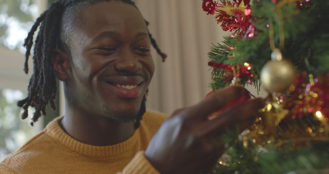 Young man smiling while placing decorations on Christmas tree. Ideal for holiday advertisements, family celebration promotions, Christmas greeting cards, and festive season campaign visuals. Emphasizes theme of joy, celebration, and traditional holiday decoration.