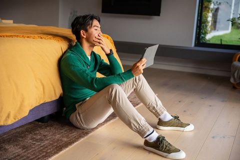 Relaxing on floor reading tablet by bed in sunlit minimalist bedroom
