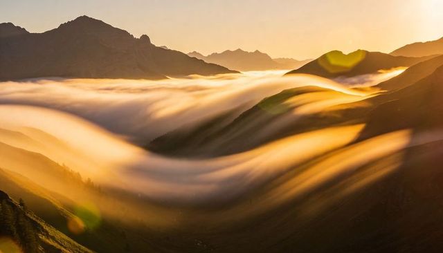 Golden fog flowing through alpine valley at sunrise, silhouetting jagged peaks