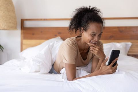 Smiling woman relaxing on bed using smartphone for morning messages and cozy home comfort