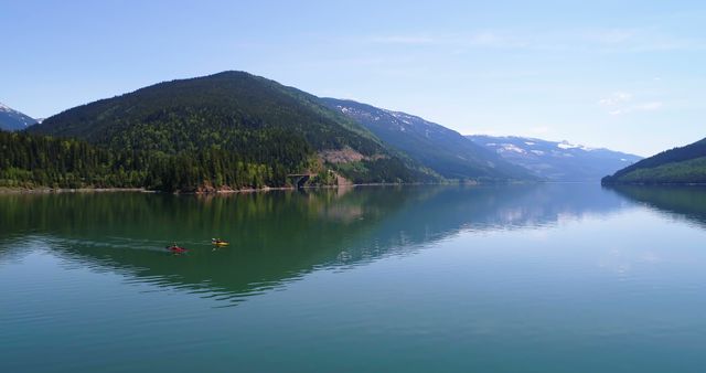 Kayakers enjoy a serene paddle on a calm mountain lake, with copy space. The tranquil waters and surrounding forested hills create a perfect setting for outdoor recreation and relaxation.