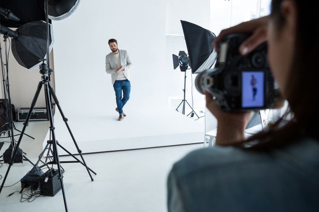 Male model posing in a professional photography studio with lighting equipment and a photographer capturing the moment. Ideal for use in articles about fashion photography, modeling careers, behind-the-scenes looks at photoshoots, and professional studio setups.