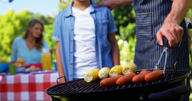 Family having barbecue in garden with person grilling hot dogs and corn on grill, child standing nearby and another adult in background enjoying time outside. Ideal for use in summer outdoor activity promotions, food blogs, family event materials, picnic gear advertisements, and lifestyle articles focusing on outdoor recreation.