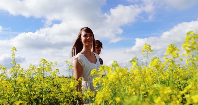 Young woman in white dress smiling while walking through a meadow filled with yellow flowers under a bright blue sky with scattered clouds. Ideal for concepts related to happiness, relaxation, nature walks, outdoor activities, summer weekends, and countryside escapes.