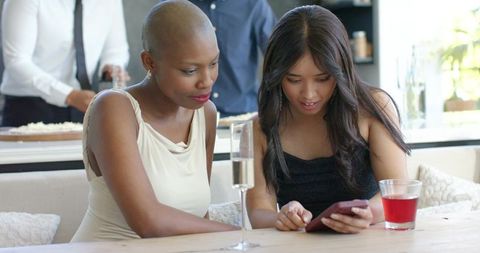 African american and asian women sharing smartphone and drinks at bright home gathering