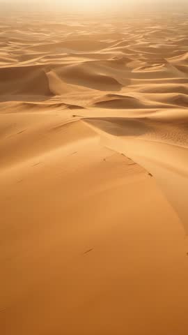 Vertical drone video revealing endless sunlit sand dunes, ripples, ridges and hazy horizon