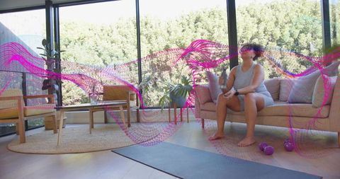 Woman resting on sofa holding phone and dumbbells after home workout in sunlit living room