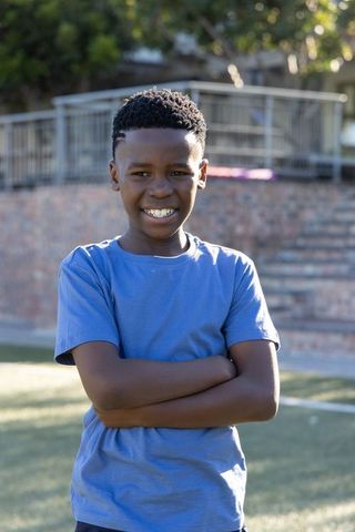 African american boy standing in sunlit park smiling with arms crossed in blue t-shirt