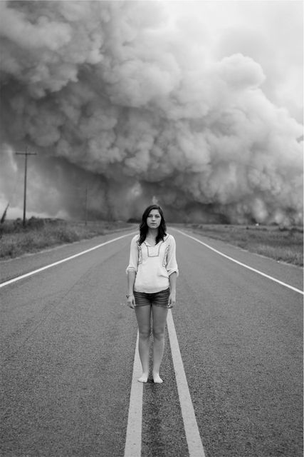 Tall woman wearing casual clothes stands barefoot on an empty rural road as a massive smoke cloud looms behind her. Her serious expression adds to the intense atmosphere of the scene. Perfect for use in art projects, blogs about environmental issues, and other creative projects needing a strong emotional impact.