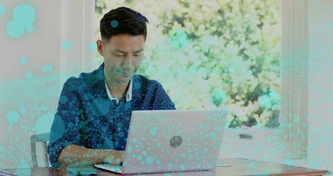 Typing Asian man working on laptop by window with turquoise bubble overlay and nature view