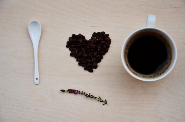 Overhead view of coffee beans arranged in heart shape next to cup of coffee, white spoon, and small flower on wooden background. Ideal for use in cafes, coffee promotions, blogs about coffee, or lifestyle articles emphasizing relaxation and enjoyment.