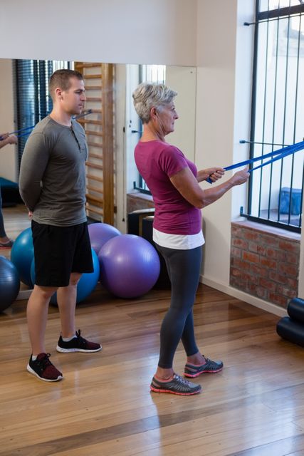 Senior woman performing resistance band exercise under the guidance of a physiotherapist in a clinic. Ideal for use in articles or advertisements related to physical therapy, senior fitness, rehabilitation programs, and healthcare services.
