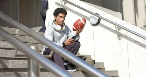 Asian college student checking phone while holding football on sunny urban campus steps