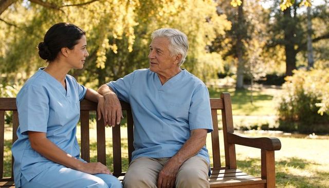 Senior man and nurse conversing on bench in sunny park offering compassionate caregiving