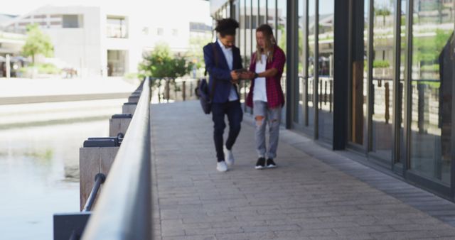 Two young professionals, one in a suit and the other in casual attire, are walking together on a paved pathway by a modern building with glass walls and a body of water in the background. They appear to be engrossed in a discussion. This image can be used in contexts related to urban work life, teamwork, modern business environments, and outdoor professional meetings.