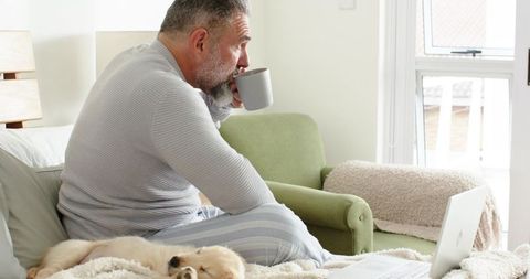 Mature man sipping coffee on bed with laptop and sleeping puppy in cozy morning