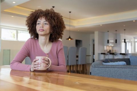 African american woman holding mug gazing out window in modern open-plan living room
