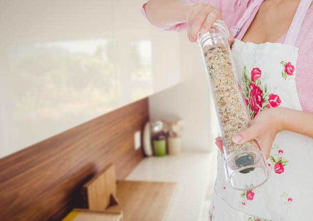 This image depicts a woman wearing a floral apron, holding a spice grinder in a modern kitchen. It is ideal for use in culinary blogs, cooking websites, and advertisements for kitchenware. Also suitable for articles related to home cooking, seasoning recipes, and modern kitchen design.