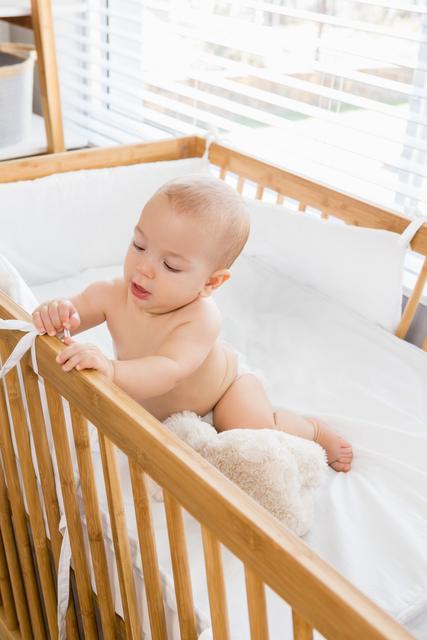 Baby boy sitting and playing in a wooden crib at home, with white bedding and bright natural light from a nearby window. Ideal for use in parenting articles, advertisements for baby products, nursery design blogs, and family lifestyle magazines.
