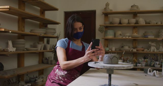 Female potter wearing apron, taking photo of her ceramic work with smartphone in pottery studio. Shelves filled with clay creations and pottery tools. Ideal for illustrating art studio environments, pottery classes, crafting processes, or social media content for artisans.