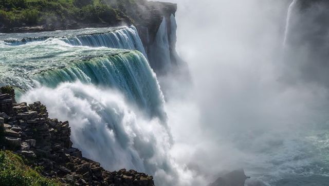 Cascading waterfall plunging into misty gorge with rocky cliff and lush green riverbank