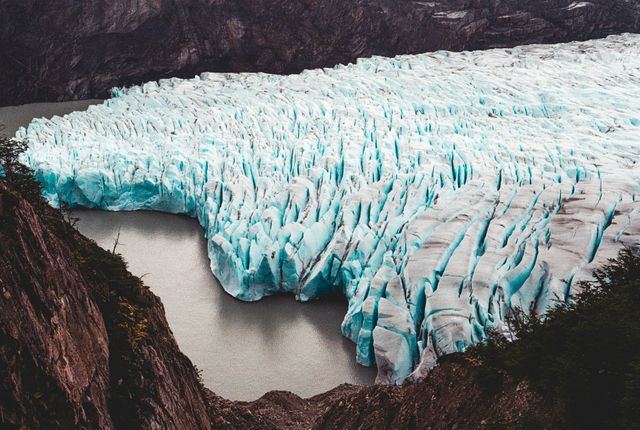 Aerial view captures stunning glacier descending into a serene mountain lake, showcasing striking ice formations and rugged terrain. Perfect for promoting outdoor travel, nature documentaries, adventure tourism, and environmental awareness campaigns.