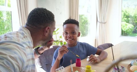 African american father painting son's cheek during easter craft at sunlit dining table