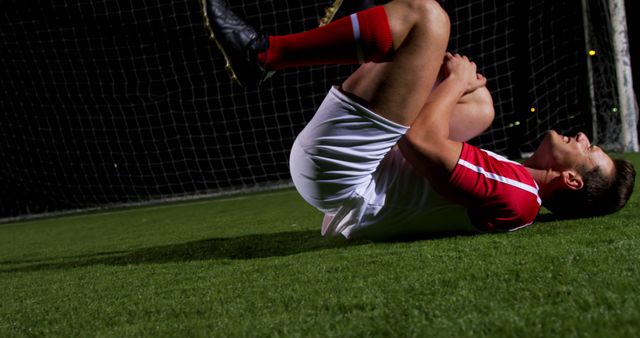 A young Caucasian male soccer player is lying on the ground, seemingly injured during a night game, with copy space. His expression and body language suggest pain and the need for medical attention on the field.
