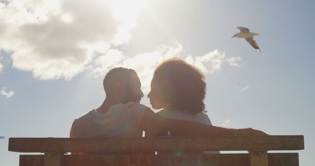 Couple is sitting on a bench, enjoying a sunny day with blue sky and white clouds while a bird flies by. Use this captured moment to depict romance, outdoor relaxation, and serene connections. Perfect for themes like love, leisure, and nature.