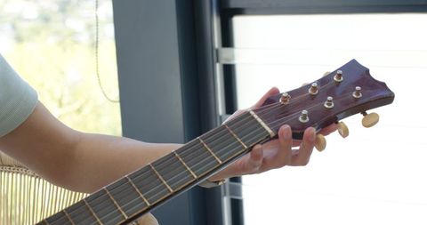 Asian female tuning acoustic guitar by window blinds adjusting tuning pegs and strings