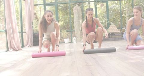 Women unrolling yoga mats in sunlit glass studio with plants for group fitness class
