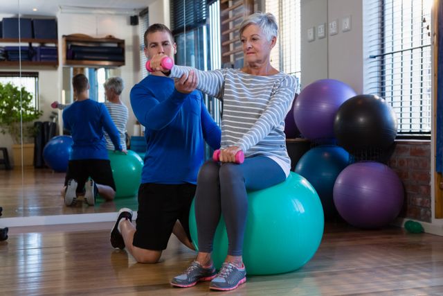 Senior woman receiving assistance from a physiotherapist while using an exercise ball and dumbbells in a clinic. Ideal for illustrating physical therapy, elderly fitness, rehabilitation programs, and health care services for seniors.
