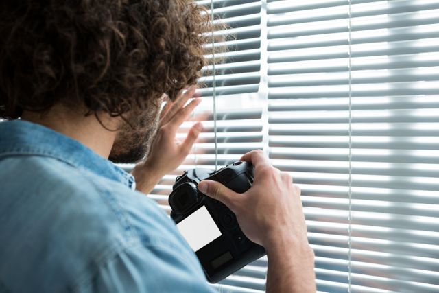 Male photographer with curly hair holding a digital camera and looking through window blinds in a studio. This image can be used for themes related to photography, creativity, professional work, and indoor studio settings. It is suitable for articles, blogs, and websites focused on photography, creative professions, and studio environments.