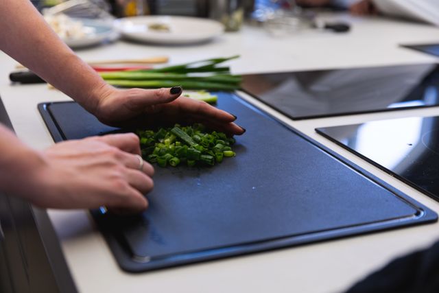 Mid section of male chef cooking in a modern busy kitchen, slicing spring onion on chopping board. Cookery class at a restaurant kitchen. Workshop cooking food.