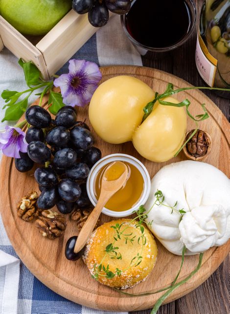 A wooden board displaying a variety of cheeses, including a ball-shaped one and fresh mozzarella, accompanied by black grapes, a small jar of honey with a wooden spoon, brown nuts, a piece of bread, and herbs. This is an ideal image for use in culinary blogs, gourmet food articles, advertisement of catering services, or restaurant menus.
