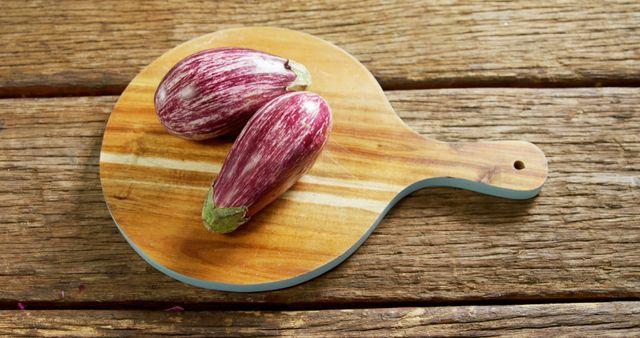 Two striped eggplants are placed on a circular wooden cutting board with handles, resting on a rustic wooden surface. The vibrant colors and rustic theme make this visual ideal for use in culinary blogs, healthy eating promotions, vegetarian recipes, and kitchen decor marketing.