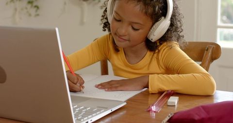 Young girl studying with laptop and headphones writing homework at kitchen table