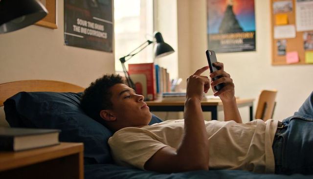 Black teen relaxing on dorm bed scrolling smartphone casual college life portrait