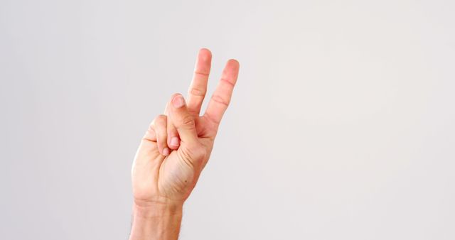 A Caucasian hand is shown making a peace sign against a plain background, with copy space. It symbolizes peace or victory and is often used in various cultural contexts.