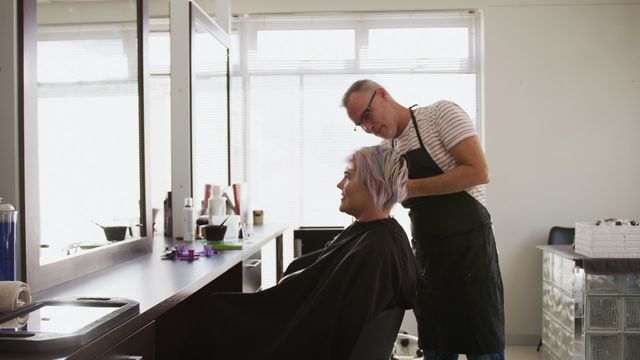 A male hairdresser styles a female client's hair in a modern salon with ample daylight creating a welcoming atmosphere. This is ideal for use in content related to beauty and fashion services, promoting friendly customer service in salons, and articles about trending hairstyles. The interaction showcases professional service and personal connection, perfect for advertisements or blogs focused on hair care or salon experiences.