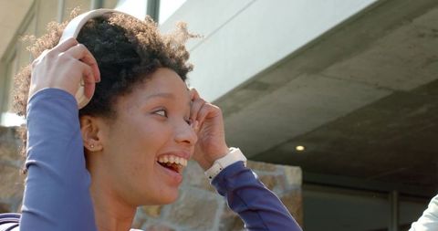 African american woman laughing adjusting headphones sunny modern campus wearing white smartwatch