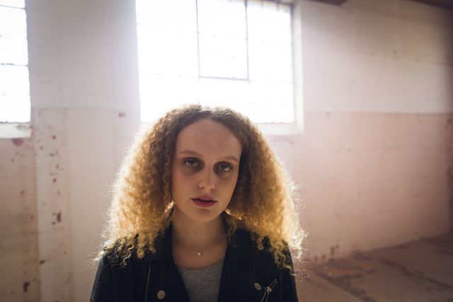 Portrait of a hip young Caucasian woman in an empty warehouse, looking straight to camera, backlit by sunlight by the window.