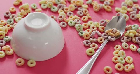 Upside-down white bowl with stainless-steel spoon and scattered pastel cereal rings on pink