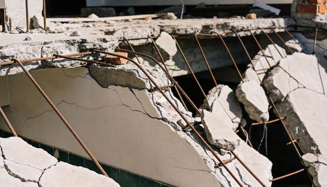 Collapsed concrete slab exposing rusted rebar, fractured plaster and dark green tile
