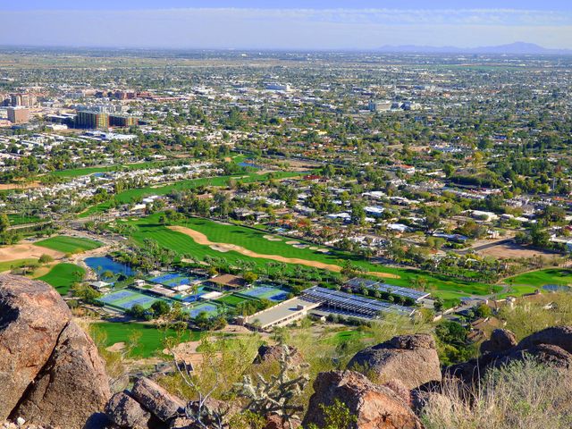 Aerial view featuring a golf course embedded within an urban setting on a sunny day. The image highlights green fields, residential buildings, and distant mountains, illustrating the coexistence of nature and city life. This photo is ideal for use in travel guides, urban planning presentations, golf course marketing materials, and articles on the blend of nature and urban development.