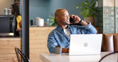 African american man working remotely in coffee shop, talking on phone, using laptop