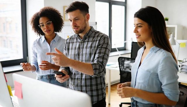 Young professionals collaborating at desk, pointing at monitor while holding smartphone